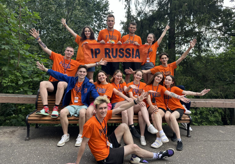 Группа студентов в языковом лагере IP Russia на природе, в оранжевых футболках, держат баннер.